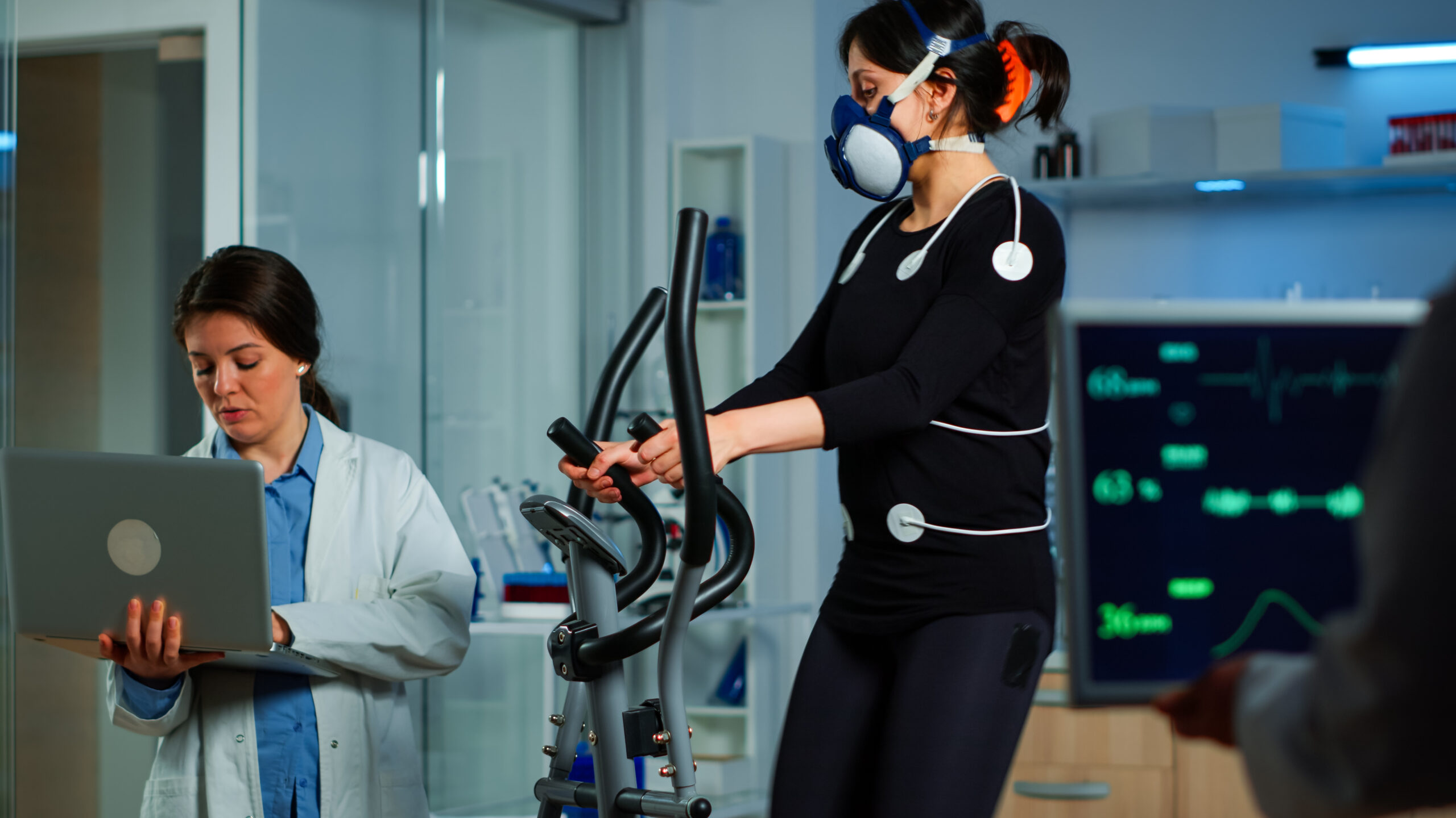 medical researcher using laptop while measuring woman endurance using body sensors, electrodes and mask measuring cardiac rhythm